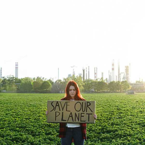 Young woman with polluting power plant in background holds a sign: "SAVE OUR PLANET"